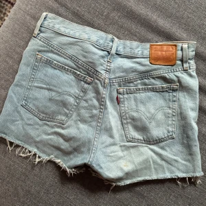 Levi's ljusblå denimshorts W28 - Säljer ett par klassiska ljusblå denimshorts från Levi's Premium. Modellen har hög midja, råa fransiga benslut och fem fickor. Knappar framtill och ikonisk läderpatch bak. Liten fläck på rumpan. 