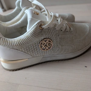 Vita sneakers från Mexx med gulddetaljer - Snygga vita sneakers från Mexx med stickad ovandel och gulddetaljer på sidor, snörning och häl. Skorna har rund tå, platt sula och är tillverkade i en mix av textil och syntet. Perfekta för dig som gillar stilrena och trendiga sneakers med lite extra bling.