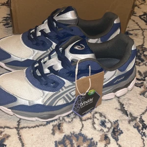 Asics blå och silver sneakers strl 44 - Snygga Asics sneakers i blått och silver med mesh och syntetpaneler. Skorna har rund tå, platt sula och snörning. Ortholite innersula för extra komfort. Perfekt för dig som gillar sportig stil och vill ha något som sticker ut.