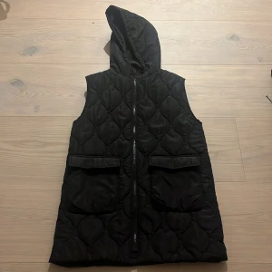 Svart  barndunväst från Zara - Svart lång barndunväst från Zara med huva och cool quiltad design. Västen har två stora fickor framtill och stängs med dragkedja. Perfekt för lager-på-lager och streetstyle-vibe.