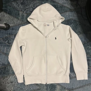 Vit Polo Ralph Lauren Zip Hoodie - PRIS KAN DISKUTERAS! Vit hoodie från Polo Ralph Lauren med klassisk broderad logga på bröstet. Säljer pga att den är för liten, De finns även snören men dem ligger inte i luvan då dem ramlat ut, så snören skickas åt sidan om tröjan. Den är XS och passar ungefär dig som är 160-170cm skulle jag chansa på!!