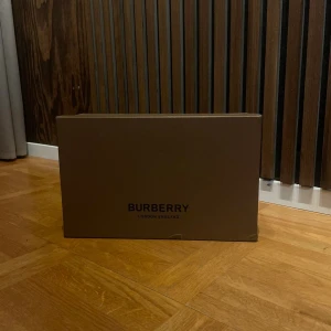 Burberry sneakers med rutig detalj - Snygga Burberry sneakers med klassisk rutig panel i beige, svart och vit. Skorna har svart skinn och mocka på ovansidan, vita platta sulor och svarta snören. Perfekt för dig som gillar stilrena och lyxiga detaljer med en modern touch.
