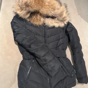 Svart hollies pufferjacka med bälte - Svart pufferjacka med stor fluffig päls på huvan och leopardmönstrat innerfoder. Jackan har dragkedja framtill, två fickor med dragkedja och ett brett elastiskt bälte i midjan för snygg passform. Perfekt för kalla dagar med sin varma och trendiga look.