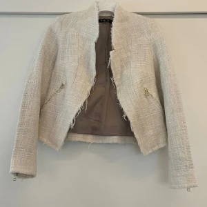 Beige kavajjacka från Mohito, strl 34 - Stilren beige kavajjacka från Mohito i storlek 34. Jackan har råa kanter, dragkedjor vid ärmslut och fickor samt en strukturerad, vävd yta som ger en exklusiv känsla. Perfekt för dig som gillar en trendig och chic look.