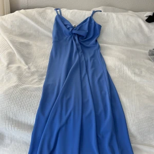 Blå långklänning med rosett XS - Supersnygg blå långklänning med smala axelband och en stor rosett framtill. Klänningen har en smockad rygg för extra komfort och sitter fint över kroppen. Materialet är mjukt och har ett lätt fall, perfekt för sommarkvällar. Passar dig som gillar stilrena och trendiga plagg. Jag är 172cm lång och den sitter lite längre ner än mina knän.