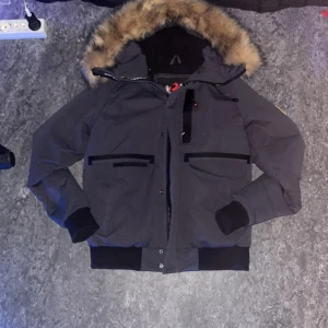 Mörkgrå dunjacka från Zavetti Canada - Säljer en mörkgrå dunjacka från Zavetti Canada med fluffig päls på huvan och snygga svarta detaljer. Jackan har flera fickor med dragkedja, ribbade muddar och ett broderat märke på ärmen. Perfekt för kalla vinterdagar och har en schysst, lite loose passform.