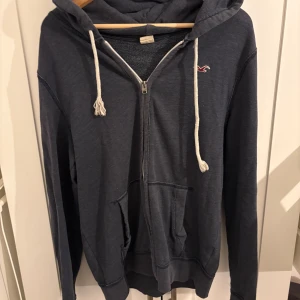 Mörkblå hoodie från Hollister - Mysig mörkblå hoodie från Hollister med vit snörning och dragkedja framtill. Tröjan har huva, två fickor och det klassiska Hollister-loggan broderad på bröstet. Perfekt för chill dagar.