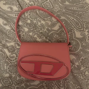 Rosa axelväska - Säljer en snygg rosa axelväska från D med ett stort metalliskt logodetalj på framsidan. Väskan har en stilren, rundad form och ett justerbart axelband. Perfekt för att lyfta din outfit med en poppig färg och cool design.