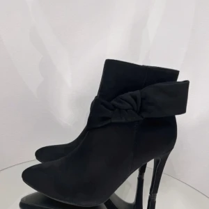 Svarta stövletter med rosett och klack - Snygga svarta stövletter med spetsig tå och hög klack. Skorna har en dekorativ rosett på sidan och är tillverkade i mocka som ger en lyxig känsla. Dragkedja på insidan för enkel på- och avtagning. Perfekta för dig som vill ha en elegant och trendig look.