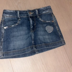 Jeanskjol med hjärtdetalj Benetton XXS barn  - Snygg kort jeanskjol från Benetton i klassisk blå denim. Kjolen har fem fickor, bälteshällor och en söt hjärtformad detalj på ena sidan. Storlek XXS barn.