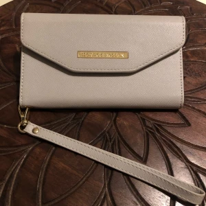 Grå/beige plånbok från Ideal of Sweden - Stilren grå/beige plånbok från Ideal of Sweden med guldfärgad logga framtill. Plånboken har avtagbar handledsrem och är tillverkad i ett syntetmaterial med lätt struktur. Perfekt för att bära det viktigaste med dig på ett snyggt sätt.