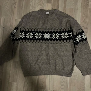 Brun stickad tröja med snöflingor - Stickad brun tröja från Lindex med klassiskt snöflingemönster i vitt och svart över bröstet och ärmarna. Rund hals och ribbade muddar vid ärmslut och nederkant. Perfekt för kalla dagar när du vill ha en mysig och stilren look.