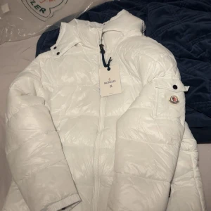 Vit Moncler pufferjacka med huva 2XL - Säljer en helt ny vit pufferjacka från Moncler i storlek 2XL Jackan har huva, dragkedja framtill och Moncler-logga på ärmen. Den är quiltad och har en glansig finish som ger en riktigt fräsch look. Perfekt för kalla dagar när du vill hålla stilen. Med NFC Scan och huva som går att dra av 