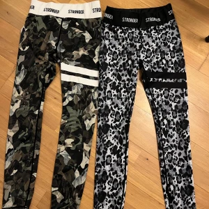 Stronger leggings med camo & leopardmönster - Två par leggings från Stronger, ett par med grönt camouflagemönster och vita ränder, ett par med svartvitt leopardmönster och svarta detaljer. Båda har hög midja och bred resår med logga. Materialet är stretchigt och består av polyester och spandex.