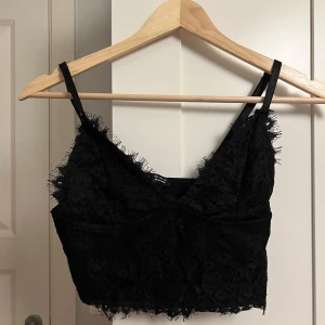 Svart spetsig croppad topp från SHEIN - Svart croppad topp från SHEIN med tunna axelband och spetsdetaljer längs kanterna. Toppen har en v-ringad form och är täckt av blommig spets framtill, vilket ger en edgy och trendig look. Perfekt för att lyfta vilken outfit som helst.