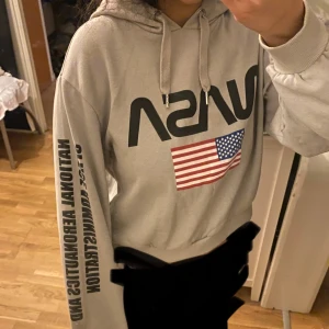Grå NASA hoodie med amerikansk flagga - Säljer en grå hoodie med NASA-tryck och amerikansk flagga. Tröjan har snörning i huvan och svart text längs ena ärmen. Strl S men passar mig som är XS