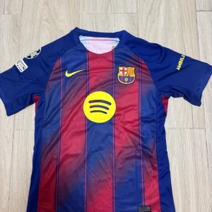 FC Barcelona Lamine Yamal matchtröja - Barça matchtröja med Lamine Yamal och nummer 10 på ryggen. Klassiska blå och röda vertikala ränder, gula detaljer och klubbmärke på bröstet. Tillverkad i lätt funktionsmaterial, kortärmad och med Champions League-märke på ärmen.