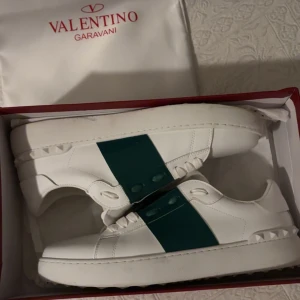 Valentino Garavani Open sneakers vit/grön - Säljer ett par snygga Valentino Garavani Open sneakers i vitt skinn med bred grön rem över mitten. Skorna har rund tå, platt sula och coola nitar på hälen. Klassisk logga i guld på plösen och vita snören. Perfekt för dig som gillar exklusiv streetstyle.