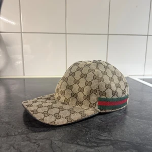 Gucci keps med GG-mönster - Säljer en beige keps från Gucci med klassiskt GG-monogram över hela kepsen. Kepsen har grönt och rött band på sidan och justerbar rem baktill i mörkbrunt skinn. Materialet är canvas och detaljerna ger en lyxig känsla. Perfekt för dig som vill sticka ut med en ikonisk accessoar.