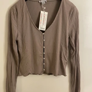 Beige ribbad blus med pärlknappar - Säljer en beige ribbad blus från Amisu  i storlek XL. Blusen har lång ärm, v-ringning och är croppad i modellen. Framtill sitter dekorativa pärlknappar som ger en extra touch. Materialet är mjukt och lätt, perfekt för dig som gillar stilrena detaljer.
