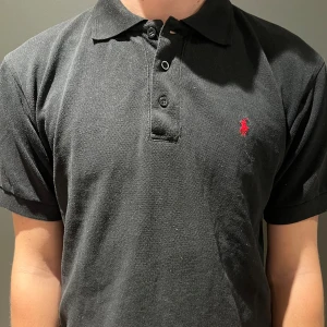 Svart pikétröja från Polo Ralph Lauren - Svart pikétröja från Polo Ralph Lauren med klassisk krage och tre knappar framtill. Tröjan har korta ärmar och den ikoniska röda Polo-loggan broderad på bröstet. Tillverkad i mjuk bomull som är skön mot huden och passar perfekt till jeans eller shorts.