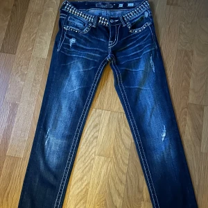 Miss Me skinny jeans med nitar, blå - Snygga blå skinny jeans från Miss Me med coola nitar och strass på linningen. Jeansen har slitningar, kontrastsömmar och klassisk femficksdesign. Märkespatch bak och detaljerade fickor ger extra edge. Perfekt för dig som gillar statement-plagg.