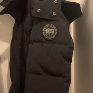 Svart dunjacka från Canada Goose S - Svart dunjacka från Canada Goose med klassisk Arctic Program-logga på ärmen. Jackan har dragkedja och tryckknappar framtill, samt detaljer i silver. Perfekt för kalla dagar med sin fluffiga och stilrena look. Snygg och tidlös design som funkar till allt.