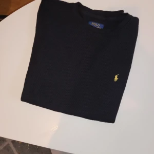 Mörkblå stickad tröja från Polo Ralph Lauren - Snygg mörkblå stickad tröja från Polo Ralph Lauren med klassisk gul broderad logga på bröstet. Tröjan har rund halsringning och långa ärmar, perfekt för lager-på-lager under kyliga dagar. I nyskick 