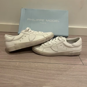 Vita Philippe Model sneakers - Snygga vita sneakers från Philippe Model med lågt skaft och klassisk snörning. Skorna har en rund tå, platt sula och är tillverkade i skinn med diskret logomärke på sidan. Perfekta för dig som gillar stilrena och trendiga sneakers.