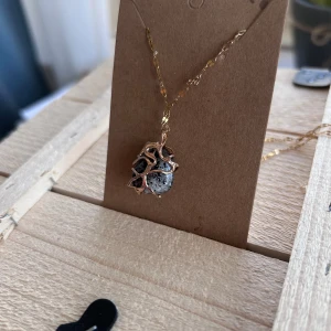 Unikt halsband med sten och guldton - Ett halsband med en kedja i guldton och ett hänge med en svart- och gråmelerad sten omsluten av ett organiskt, grenliknande mönster i guld. Kedjan är tunn och glansig, och hänget ger ett coolt och konstnärligt intryck. Perfekt för dig som gillar unika smycken.