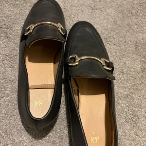 Svarta loafers med gulddetalj H&M - Stilrena svarta loafers från H&M med snygg guldfärgad metallspänne över foten. Skorna har en klassisk rund tå och är gjorda i ett slätt syntetmaterial med beige innersula. Perfekta för en clean och trendig look.