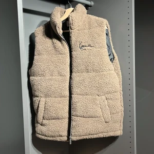 Beige teddyväst från Karl Kani - Mysig beige teddyväst från Karl Kani med broderad logga på bröstet. Västen har hög krage, två fickor framtill och stängs med dragkedja. Perfekt för lager-på-lager och ger en cool streetstyle-vibe.