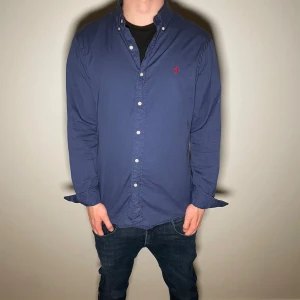 Blå skjorta Ralph Lauren - Marinblå skjorta från Ralph Lauren med button-down krage och röd broderad logga på bröstet. Skjortan har långa ärmar, vita knappar och är tillverkad i mjuk bomull. Perfekt för en clean och stilren look.