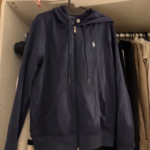 Marinblå hoodie från Polo Ralph Lauren - Snygg marinblå hoodie från Polo Ralph Lauren med dragkedja framtill, huva med snörning och klassisk logga på bröstet. Tröjan har känguruficka och är tillverkad i mjuk bomull, perfekt för chill dagar eller när du vill vara casual men ändå stilren.