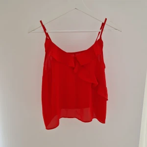 Röd volang linnetopp med smala axelband - Säljer en röd linnetopp med smala justerbara axelband och snygga volangdetaljer framtill. Toppen är lätt och luftig med transparent känsla, perfekt för varma dagar. Passformen är normal och toppen har en feminin och trendig look.