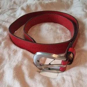 Rött skärp i äkta skinn 80 cm - Snyggt rött bälte i 100% äkta skinn med ett silverfärgat metallspänne. Bältet har en enkel och stilren design. Märkt med svensk tillverkning och har en längd på 80 cm.