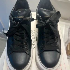 Alexander McQueen svarta sneakers - Snygga svarta sneakers från Alexander McQueen med vit plattformssula och rund tå. Skorna har breda svarta snören och guldfärgad logga på plösen. Tillverkade i skinn med stilren design och chunky siluett som ger en cool vibe.