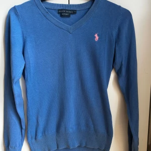 Blå v-ringad tröja från Ralph Lauren - Snygg blå v-ringad tröja från Ralph Lauren med rosa logga på bröstet. Tröjan har lång ärm, ribbade muddar och är i en tunn stickad bomullskvalitet. Perfekt för lager-på-lager. skriv om ni vill ha fler bilder på tröjan eller om ni vill se den på!🫶skulle säga att de är en s/xs