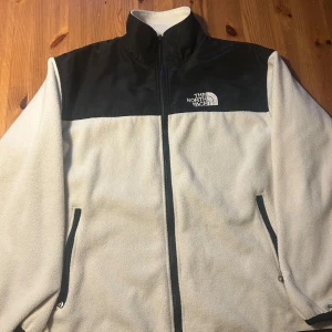 Vit fleecejacka The North Face - Säljer en snygg fleecejacka från The North Face i vit och svart. Jackan har hög krage, hel dragkedja och två sidofickor med dragkedja. Överdel i svart och resten i vit fleece, perfekt för kyliga dagar. Logga broderad på bröstet.