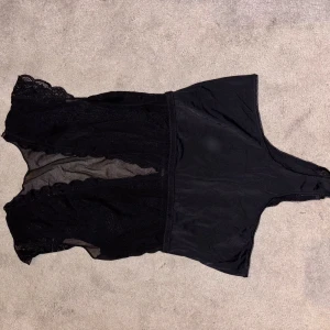 Svart spetsig body  - Säljer en svart body med snygga spetsdetaljer och transparenta partier framtill. Toppen har en ärmlös design och markerad midja, perfekt för att skapa en edgy look. Materialet är en mix av spets och syntet som ger en cool kontrast.