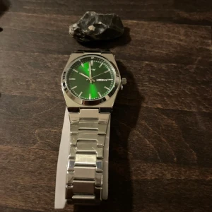 Grön och silverfärgad TPW klocka - Skriv gärna vilken färg ni vill ha innan Snygg klocka från TPW med grön urtavla och dag/datumvisning. Boetten och länken är i blankt silverfärgat metall, vilket ger en clean och modern look. Klockan har en klassisk rund form och stilrena indexmarkeringar. Perfekt för dig som gillar stilrena accessoarer.