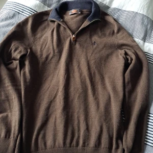 Brun stickad half zip-tröja Morris - Snygg brun stickad tröja från Morris med mörkblå krage och half zip-dragkedja. Broderad logga på bröstet och diskret emblem på ryggen. Perfekt för lager-på-lager och chill höst- och vinterstil.