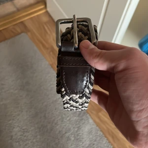 Flätat brunt och beige bälte - Snyggt flätat bälte i brunt, beige och svart med detaljer i mörkbrunt skinn och silverspänne. Bältet har en cool, vävd look som passar perfekt till jeans eller chinos. Ger outfiten en avslappnad men ändå stilren känsla.