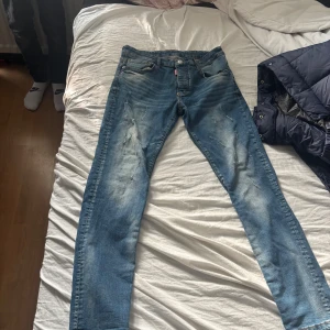 Blå Dsquared2 jeans, storlek 50 - Snygga blå jeans från Dsquared2 med slitna detaljer och klassisk femficksdesign. Jeansen har en brun läderpatch bak i midjan och orange kontrastsömmar. Passformen är rak med något smalare ben, perfekt för en trendig streetstil.