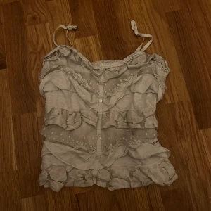 Vit volangig linnetopp Abercrombie - Supersöt vit linnetopp från Abercrombie & Fitch med volanger, spetsdetaljer och knappar framtill. Justerbara axelband och transparenta partier med broderade prickar ger en romantisk vibe. Perfekt för sommardagar när du vill känna dig extra fin.