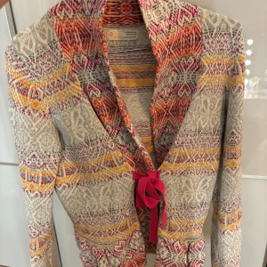 Mönstrad kofta med rosa knytband - Unik kofta från Odd Molly med färgstarkt mönster i grått, rött, gult och orange. Koftan har lång ärm och ett snyggt rosa knytband framtill. Materialet är stickat och ger en skön känsla. Perfekt för dig som gillar detaljer och färg.