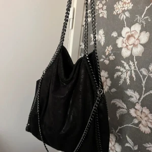 Svart axelväska med kedjedetaljer - Cool svart axelväska med grova silverfärgade kedjor som axelremmar och längs kanterna. Väskan är mjuk och har en lätt glansig yta, troligen i syntet eller mockaimitation. Rymlig insida med dragkedjefack. Perfekt för dig som gillar edgy och trendig stil.