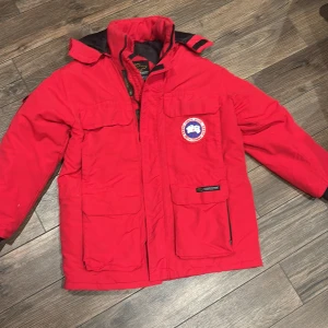  Canada Goose L - Säljer en röd Canada Goose Expedition dunjacka i storlek L. Jackan har flera stora fickor, huva och klassisk patch på bröstet. Tillverkad i slitstark nylon med fyllning av ankdun, perfekt för kalla vinterdagar. Snygg och funktionell design med dragkedja och kardborre vid ärmslut.