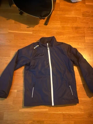 Mörkblå jacka från CCM  - Snygg mörkblå vindjacka från CCM i storlek XL Youth. Jackan har vit dragkedja framtill och två sidofickor med dragkedja. Lätt och sportig design med högt krage och CCM-logga på axeln. Perfekt för dig som gillar en clean och sportig look.