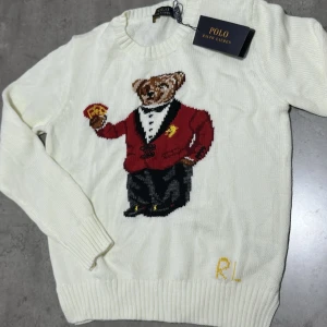 Vit stickad tröja Polo Ralph Lauren - Vit stickad tröja från Polo Ralph Lauren med lång ärm och rund hals. Framsidan har en stor broderad nalle i röd kavaj och svarta byxor, samt RL-broderi i gult nedtill. Tröjan har ribbade muddar och är mjuk i materialet.